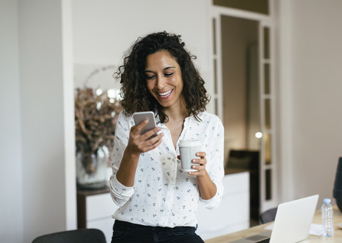 Geschäftsfrau im Büro, die ein Smartphone benutzt und eine Tasse Kaffee in der Hand hält, lizenzfreies Stockfoto