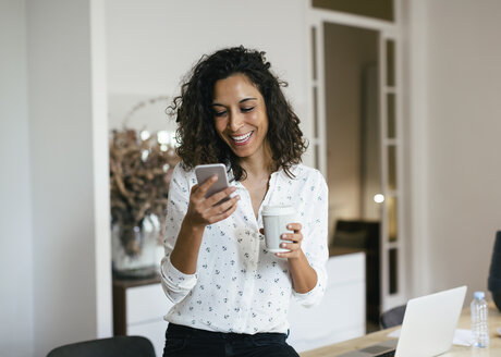 Geschäftsfrau im Büro, die ein Smartphone benutzt und eine Tasse Kaffee in der Hand hält - EBSF01936