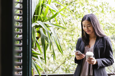 Young woman standing at window reading messages - EBSF01939