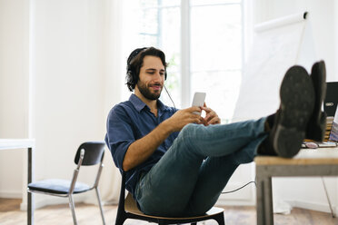 Businessman in office using headphones and smart phone, feet up - EBSF01945