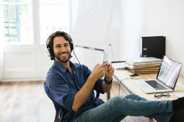 Businessman in office using headphones and smart phone, feet up - EBSF01946