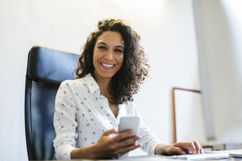Erfolgreiche Geschäftsfrau sitzt im Büro und hält ein Smartphone in der Hand - EBSF01960