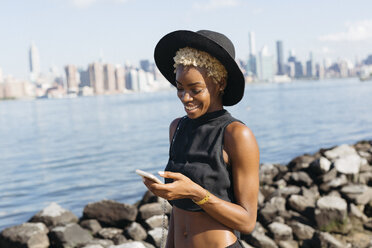 USA, New York City, Brooklyn, lächelnde junge Frau am East River, die auf ihr Mobiltelefon schaut - GIOF01647