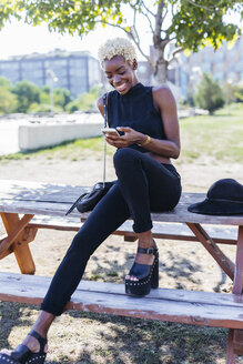 Lächelnde junge Frau sitzt auf einem Holztisch und schaut auf ihr Handy - GIOF01654
