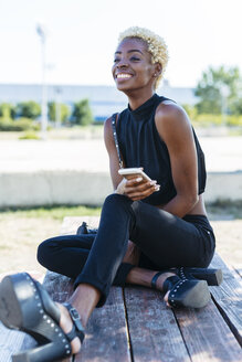 Lächelnde junge Frau sitzt auf einer Bank und hält ein Handy in der Hand - GIOF01659