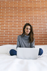 Junge Frau sitzt auf dem Bett und benutzt einen Laptop - VABF00860