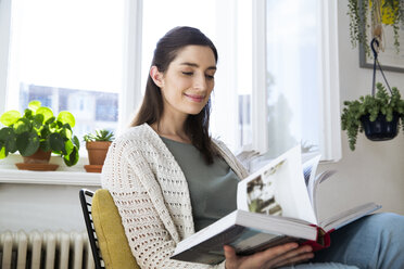 Smiling woman at home sitting on chair reading book - FKF02123