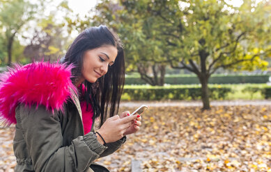 Junge Frau schaut in einem Park im Herbst auf ihr Handy - MGOF02652