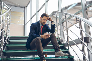 Nachdenklicher Geschäftsmann sitzt auf einer Treppe und schaut auf sein Handy - ZEDF00450