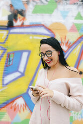 Portrait of happy young woman with earphones and cell phone in front of graffiti wall - GEMF01290