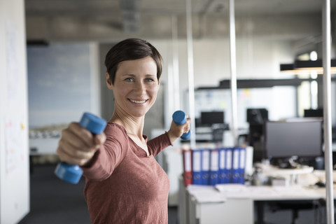 Smiling businesswoman in office exercising with dumbbells stock photo