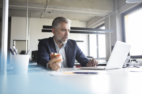 Geschäftsmann, der einen Laptop benutzt und einen Apfel am Bürotisch isst - RBF05307