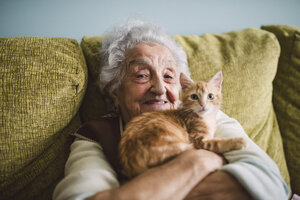 Portrait of happy senior woman cuddling with her cat on the couch - RAEF01594