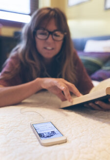 Frau mit Mobiltelefon liest zu Hause ein Buch - MGOF02678