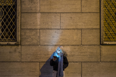 Junge Frau lehnt nachts an einer Fassade und schaut auf ihr Smartphone, lizenzfreies Stockfoto