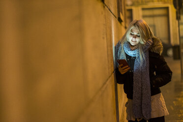 Young woman leaning against facade at night looking at her smartphone - SIPF01146