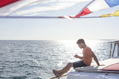 Junger Mann sitzt auf einem Boot und schaut auf sein Handy - WESTF22260