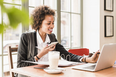 Smiling young woman using cell phone and laptop in a cafe - UUF09450