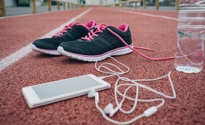 Sportschuhe, Smartphone mit Kopfhörern und eine Flasche Wasser auf der Tartanbahn - DAPF00511