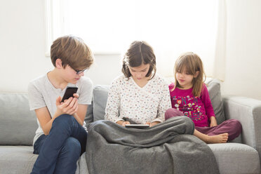 Mädchen sitzt auf der Couch und benutzt ein Mini-Tablet, während Bruder und Schwester sie beobachten - LVF05674