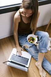 Woman at home eating vegetables with chopsticks and using laptop - VABF00897