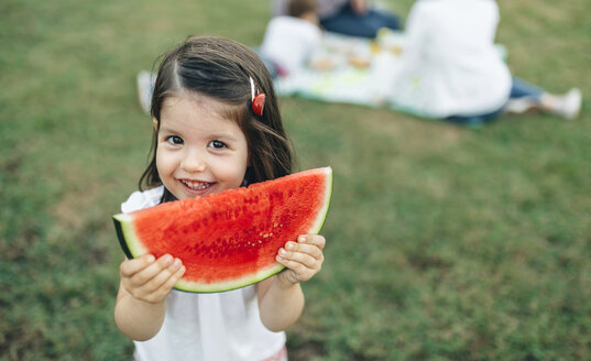 Porträt eines lächelnden Mädchens, das eine Wassermelonenscheibe hält, mit ihrer Familie im Hintergrund - DAPF00522