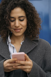 Lächelnde junge Frau mit Kopfhörern, die auf ihr Mobiltelefon schaut - KKAF00171