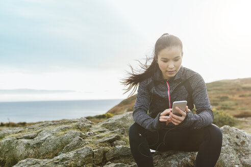 Ireland, Howth, female athlete sitting at cliff coast looking at cell phone - MADF01297