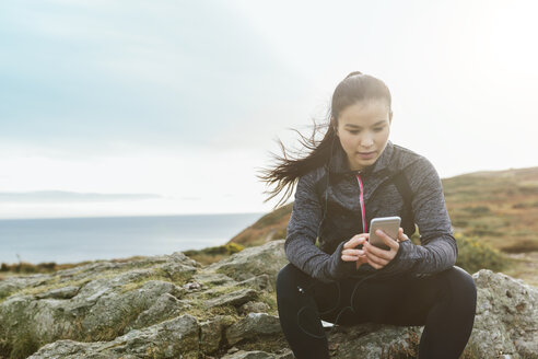 Irland, Howth, weibliche Athletin sitzt an der Klippenküste und schaut auf ihr Handy - MADF01297