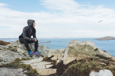 Ireland, Howth, female athlete sitting at cliff coast - MADF01303