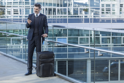 Businessman with cell phone standing with suitcase on bridge stock photo