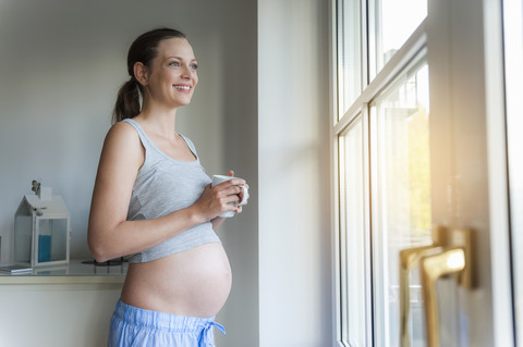 Lächelnde schwangere Frau mit Tasse am Fenster, lizenzfreies Stockfoto