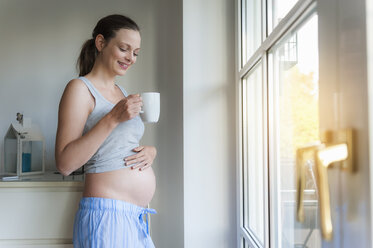 Smiling pregnant woman holding cup at the window - DIGF01501