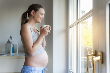 Smiling pregnant woman holding cup at the window - DIGF01502