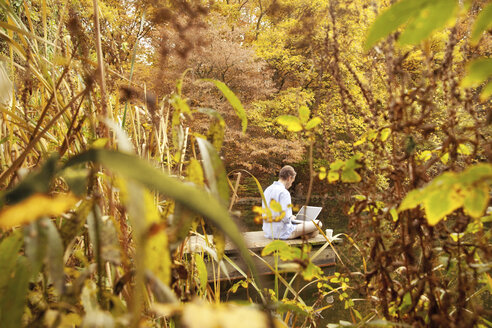 Mann mit Laptop auf einem Steg an einem Teich im Herbst - MFRF00834