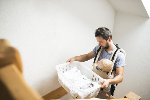 Vater mit Baby in der Babytrage, der einen Wäschekorb trägt und die Treppe hinaufgeht - HAPF01230