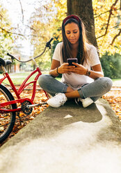 Junge Frau schaut in einem Park im Herbst auf ihr Handy - MGOF02718