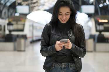 Glückliche junge Frau mit Mobiltelefon am Bahnhof - KKAF00186