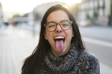Porträt einer glücklichen jungen Frau mit Brille, die ihre Zunge herausstreckt - KKAF00197