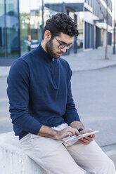 Junger Mann sitzt auf einem Poller und benutzt ein Mini-Tablet - TCF05250