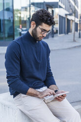 Young man sitting on bollard using mini tablet - TCF05250