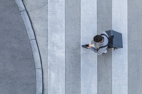 Businessman with baggage crossing the street while looking at cell phone, top view - TCF05251