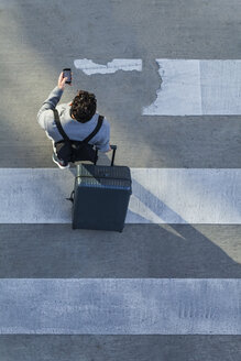 Businessman with baggage crossing the street while looking at cell phone, top view - TCF05253