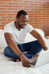 Lächelnder junger Mann mit Kopfhörern, der auf dem Bett sitzt und einen Laptop benutzt - VABF00932