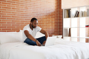 Young man with earphones sitting on bed using laptop - VABF00933