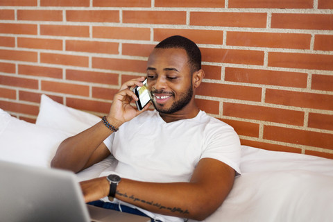 Lächelnder junger Mann auf dem Bett mit Laptop und Mobiltelefon, lizenzfreies Stockfoto