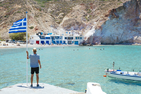 Griechenland, Milos, Firopotamos Beach, Mann schaut aufs Meer, hält sich an der griechischen Flagge fest - GEMF01331