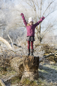 Porträt eines glücklichen Mädchens mit erhobenen Armen auf einem Baumstamm im Winter stehend - SARF03098