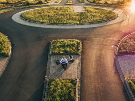 Businessman with laptop sitting on traffic island in roundabout - KNSF00813