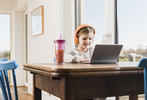 Kleines Mädchen sitzt zu Hause mit Laptop und Kopfhörern - UUF09529